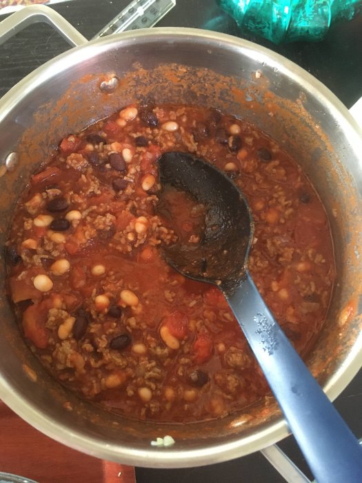 En gryta med chili con carne och en svart slev. Kokning. Böner och kött synliga. Röd sås. Hemlagat.