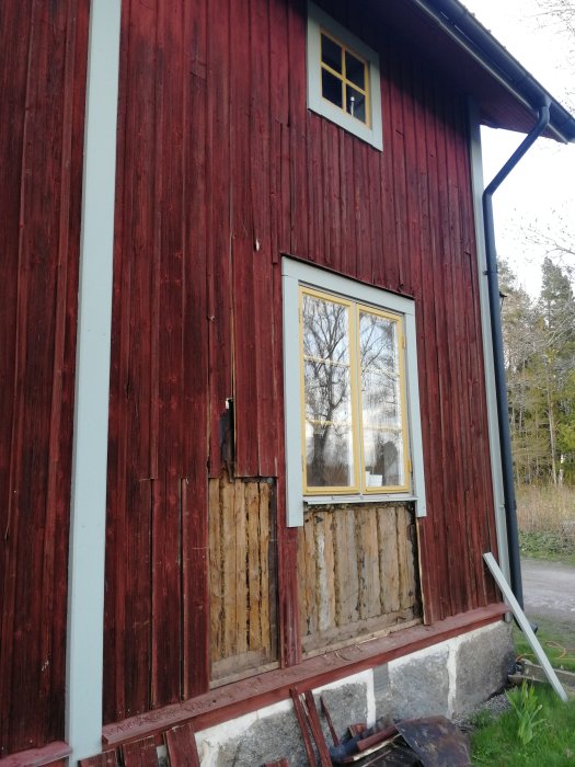 Rött trähus med skadad fasad visar underliggande material, två fönster, inga personer, renoveringsbehov, dagtid, ute.