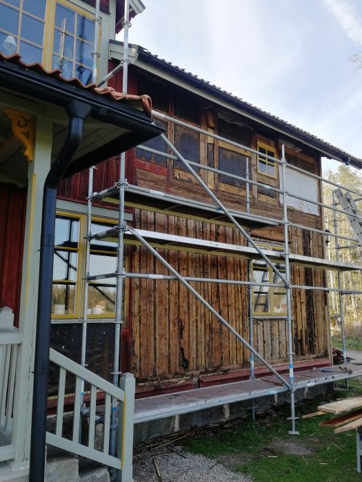 Gammalt hus under renovering med byggställningar, avskalad träfasad, fönster och grönskande omgivning.