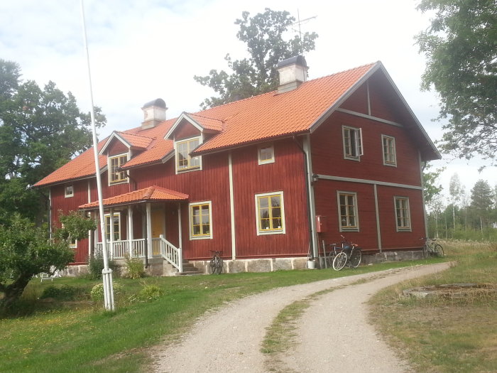 Rött trähus med vita knutar, veranda, gult fönster, grönt gräs, grusväg, cyklar, traditionell svensk landsbygdsarkitektur.