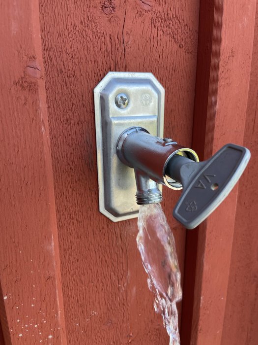 Nyckel i vattnutkast, röd vägg, vatten strömmar, metallplatta, skruvar, silverfärgad nyckel, brun handtag.