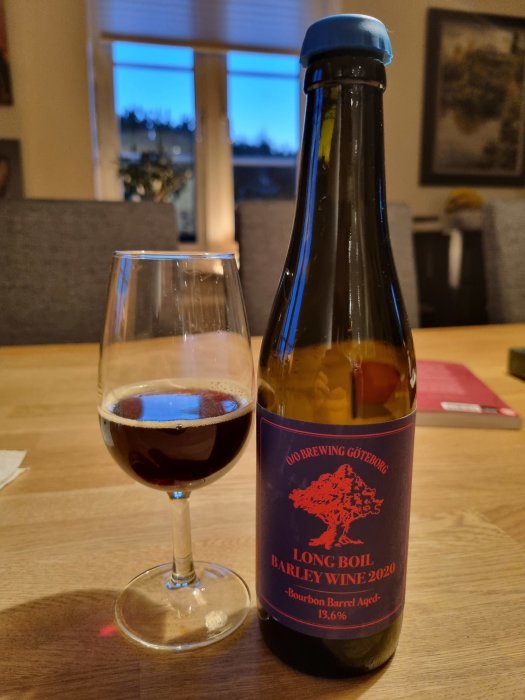 Ölflaska med etikett "Long Boil Barley Wine", glas med mörkt öl, inomhusmiljö, skymning utanför fönstret.