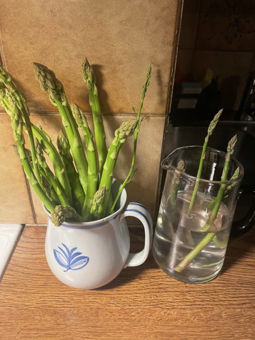 Färsk sparris i en kanna och ett glas med vatten på ett träbord.