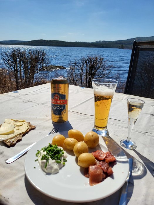 Måltid utomhus med potatis, sill, gräddfil, öl och knäckebröd vid vatten på solig dag.