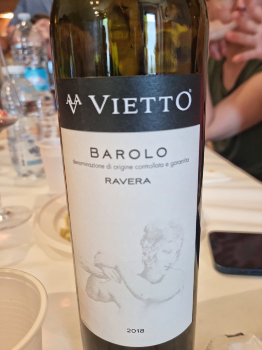 Flaska Vietti Barolo Ravera 2018 vin, etikett med konstverk, middagsmiljö, oskärpa i bakgrunden, person.