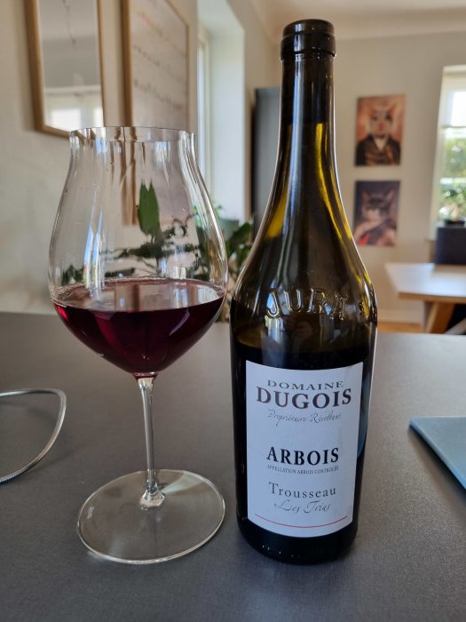 Vinflaska märkt "Domaine Dugois, Arbois Trousseau" bredvid vinglas med rött vin, inomhusmiljö.