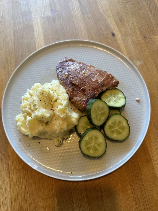 Stekt köttbit, potatismos, skivad gurka på en tallrik mot träbordsbakgrund.