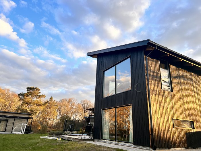 Modernt hus med stora fönster, reflektion av träden, blå himmel, moln, aftonsol, trädgård.