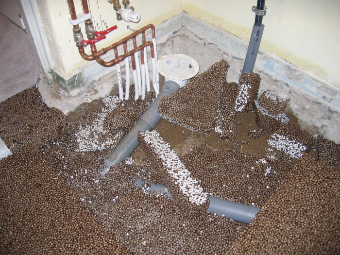 Vattenledningar, avloppsrör, grus och expanderad polystyren (frigolitkulor) på ett öppet golv.