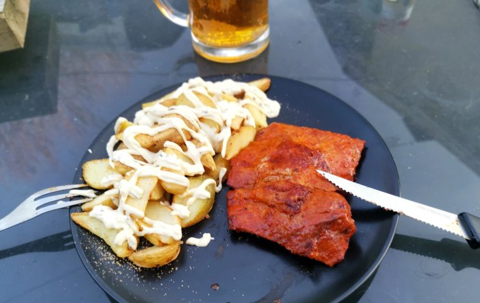 Middag utomhus: kryddig köttbit, pommes med sås på svart tallrik, ölglas i bakgrunden.