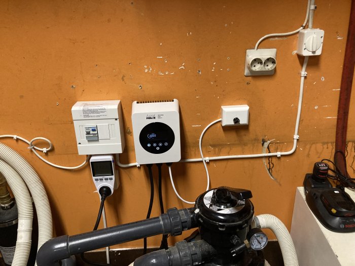 Tekniskt rum med väggmonterade elektriska apparater, kabeldragningar, rör och pumpsystem mot orange bakgrund.