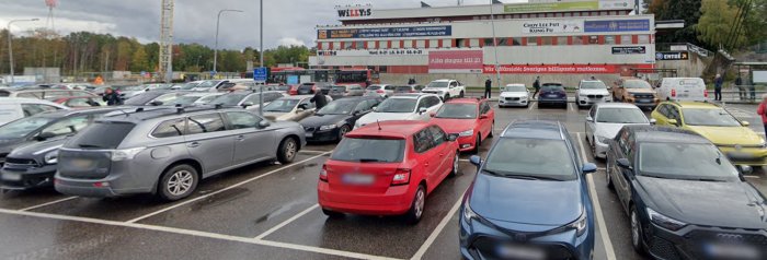 Full parkeringsplats utanför köpcentrum på molnig dag. Skyltar visar butiker och tjänster. Människor syns i bakgrunden.