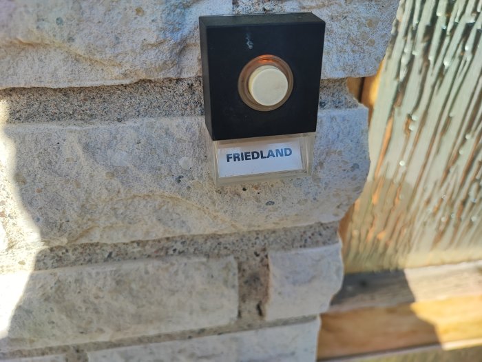 Svart dörrklocka monterad på stenmur med märkning "FRIEDLAND" i soligt väder.