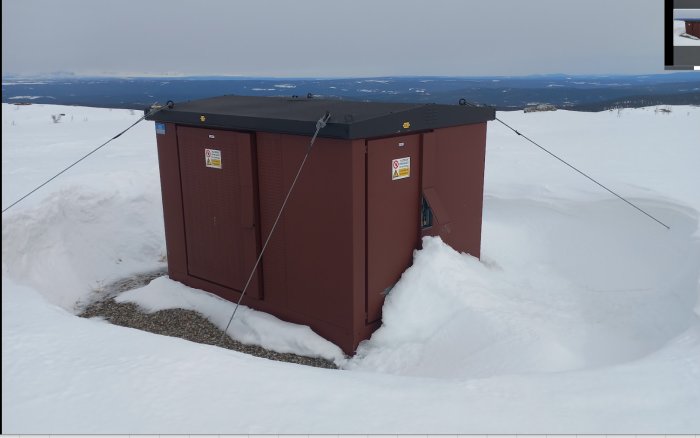 Röd byggnad med vajrar i snöigt landskap, berg i bakgrunden, grå himmel.