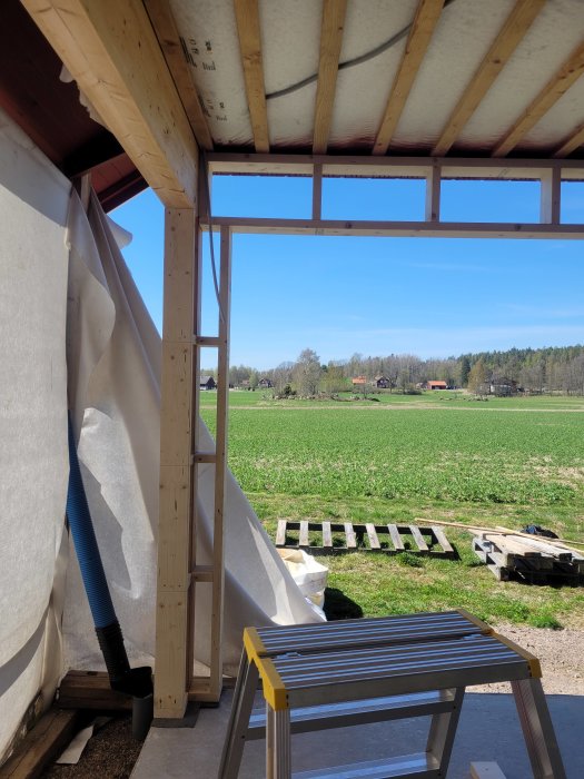 Byggarbetsplats med trästomme, isolering och plast, utsikt över fält, blå himmel, vårdag, några pallar utomhus.