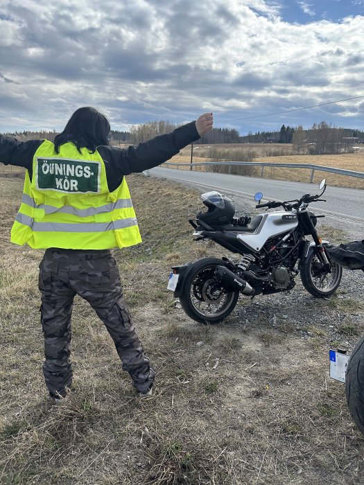 Person med "ÖVNINGSKÖR"-väst gör stopptecken vid motorcykel vid vägkant. Landskap med träd och moln i bakgrunden.