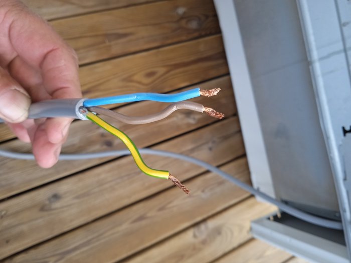 En hand håller elledningar med avskalade ändar vid en grå elcentral, osäker installation, risk för elektrisk stöt.