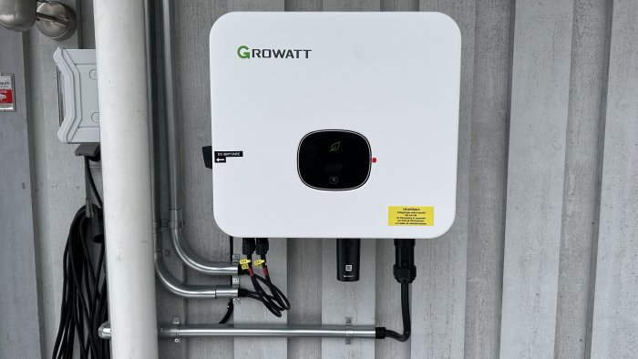 Vit Growatt solenergi inverter monterad på betongvägg, omgiven av elektriska installationer och rör.