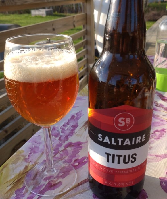 Ett glas öl och en ölflaska utomhus på en blommig duk. Brittisk ale, "Saltaire Titus", alkoholhalt 3,9%.