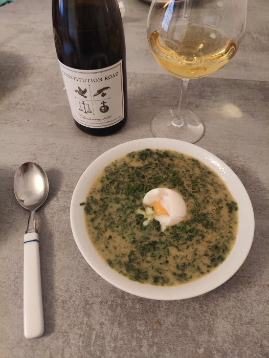 Grön soppa med pocherat ägg, flaskhals syns, vin i glas, sked, stenbord.