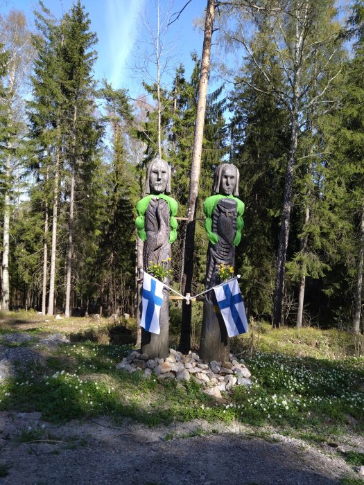 Två trästatyer klädda i grönt, håller i finländska flaggor, omgivna av skog.