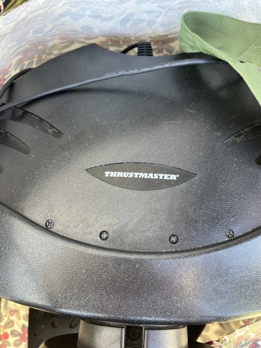 Svart ratt med märket "Thrustmaster", delvis skymd av grön tygkant, närbild, detaljerad textur.