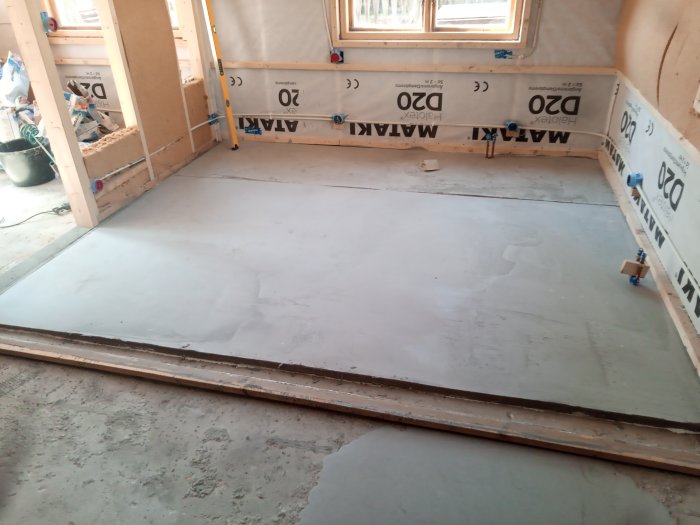 Rum under renovering, nygjuten betonggolv, isoleringsmaterial på väggarna, byggmaterial, och arbetsverktyg synliga.