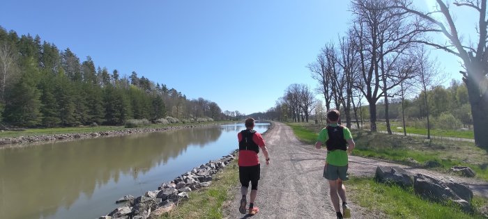 Två löpare på grusväg bredvid en stilla kanal, omgiven av träd och blå himmel.