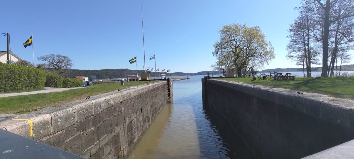 Sluss i soligt landskap, svenska flaggor, vatten, träd, blå himmel, cyklar, bänkar och lugn atmosfär.