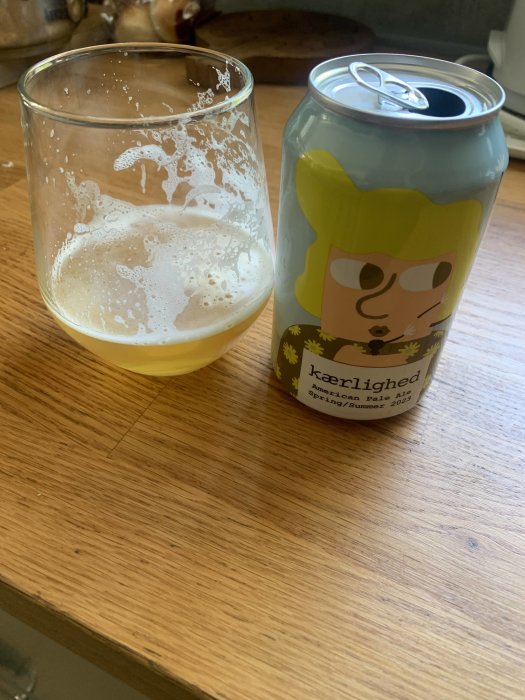 Halvtomt ölglas bredvid öppnad burk med tecknad figur, på träbord. "American Pale Ale", sommar/vår-tema.