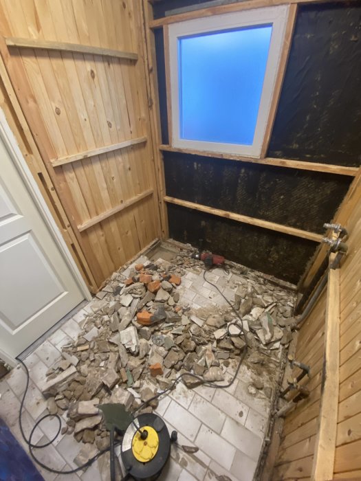 Renovering pågår, röran av tegel och kakel på golvet, träpanel vägg, fönster, dammsugare, synlig isolering.