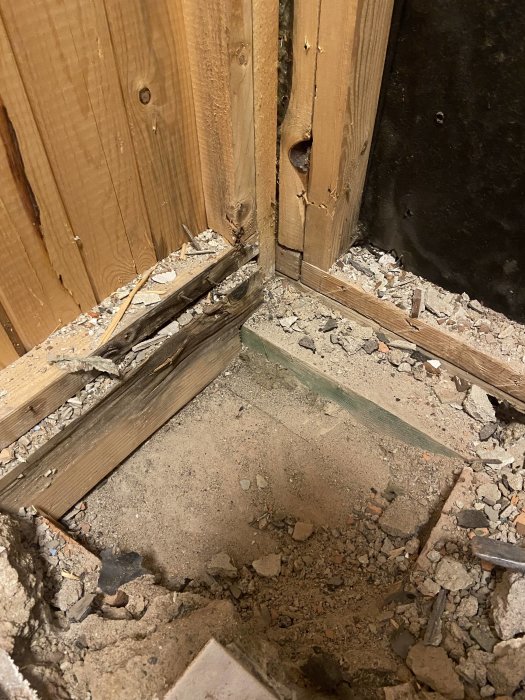 Skadade trästolpar och sönderbruten betonggrund i hörn av byggnad, tyder på reparationer eller skadedjursangrepp.