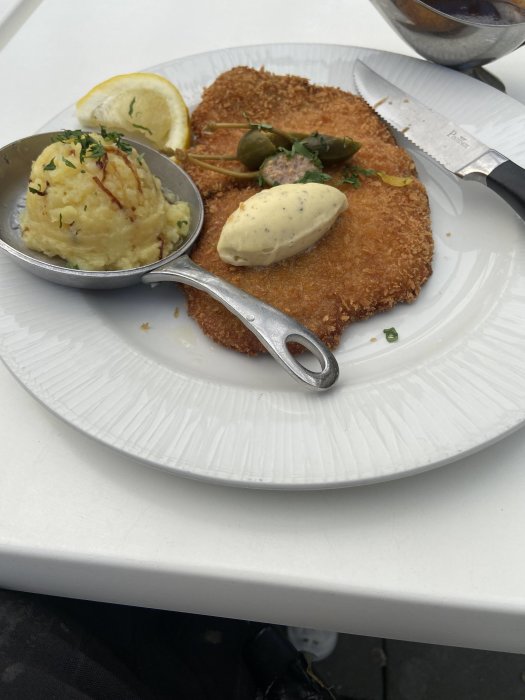 Ett panerat schnitzel på tallrik med potatismos, citron, oliver, kapris och örtsmör.