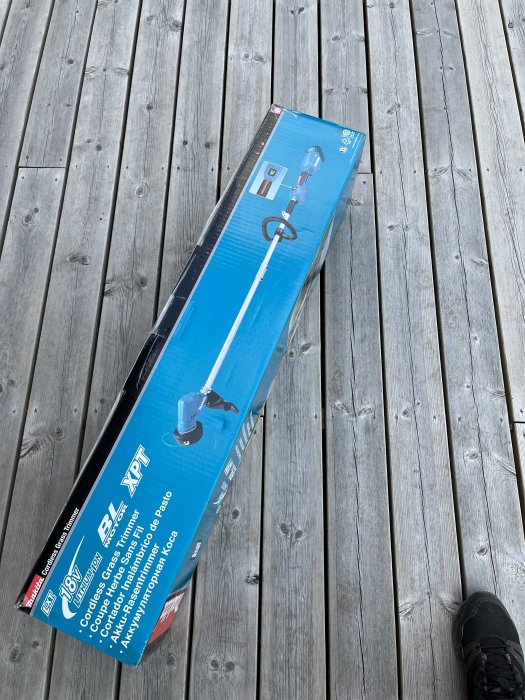 En låda med ett trådlöst grästrimmer på ett trägolv, del av en sko syns.