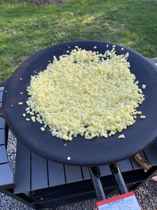 Stor svart stekhäll utomhus fylld med hackad potatis, gräsmatta i bakgrunden, matlagning utomhus.