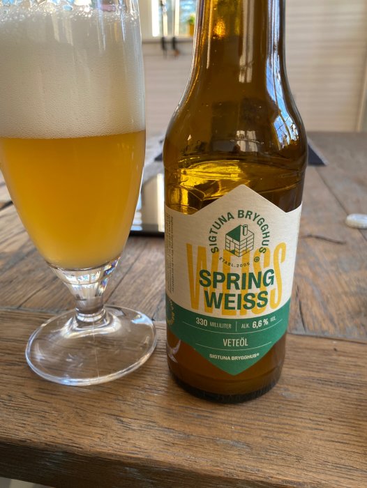 Ölglas med skum, delvis tom ölflaska, "Spring Weiss", träbord, utomhusmiljö.