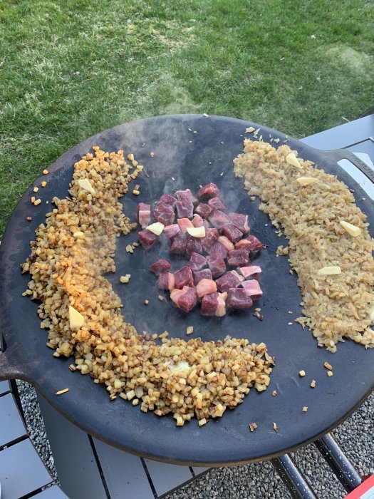 Matlagning utomhus på stor cirkulär platta, friterat ris och råa köttbitar, upplagt, smörbitar, gräs i bakgrunden.