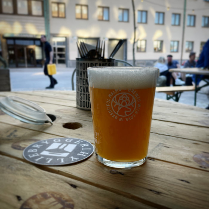 Ölglas på träbord, ölunderlägg, oskarp bakgrund, utomhusmiljö, personer sitter vid andra bord.