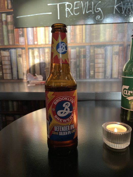 En ölflaska från Brooklyn Brewery på ett bord, bredvid ett tändljus, med bokhylla i bakgrunden.