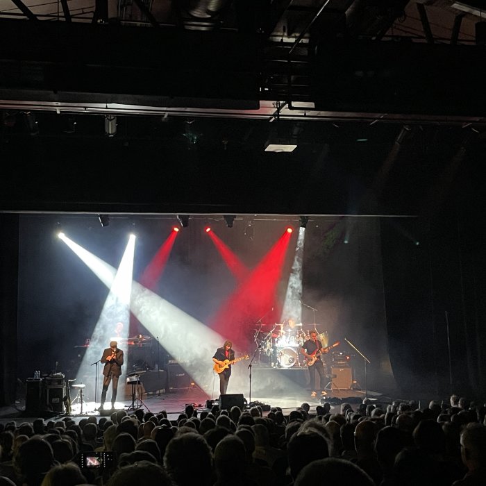 Livekonsert inomhus, scen med musiker, strålkastarljus, publik i silhuett.