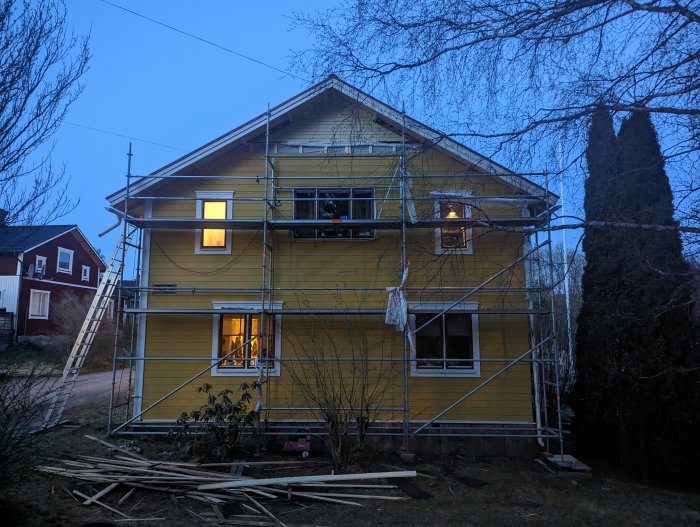 Gult hus med byggnadsställning, kvällstid, träd, skymning, byggmaterial på marken, bostadsområde.