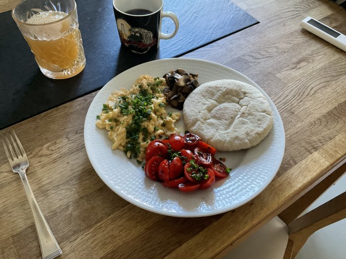 Frukost med äggröra, skivad tomat, champinjoner, pitabröd, juice och kaffe, serverad på ett träbord.