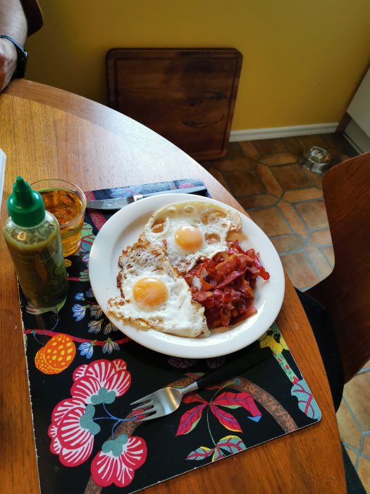 Frukosttallrik med ägg och bacon, färgglad bordstablett, trästol, gul vägg, del av person.