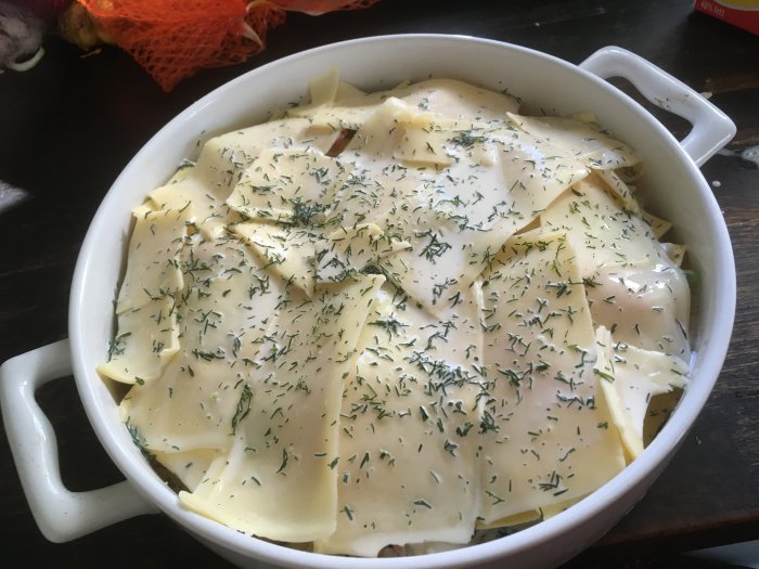 Gratäng i tillagningsfas med pastaplattor, ostsås, dill, vit bakgrundsföremål, träbord, råvarumenyn synlig.