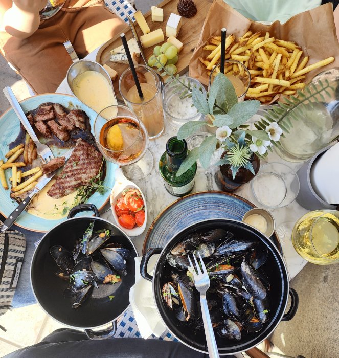 En måltid ute med musslor, steak, pommes frites, ost, vindruvor, och drycker på ett bord.