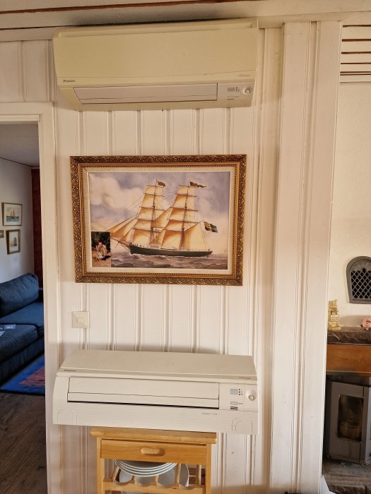Vitmålat rum med tavla av skepp, luftkonditioneringsenheter och trästol. Vintage och maritimt tema.