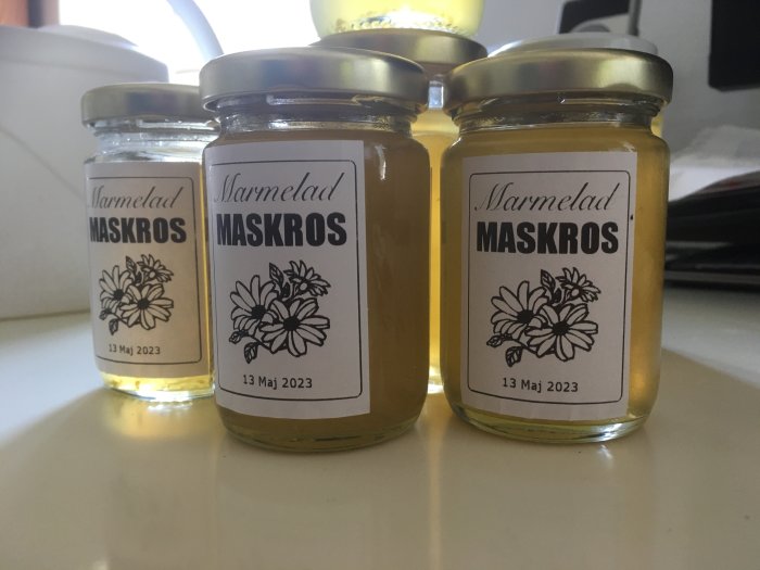 Tre burkar marmelad med maskros, etiketter märkta "13 Maj 2023", gul innehåll, köksbakgrund.