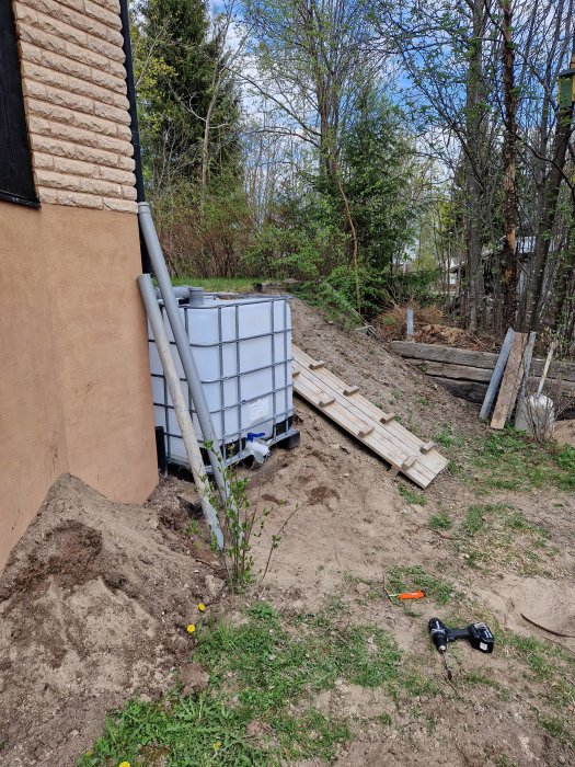 Trädgård under konstruktion, IBC-tank, bräder, verktyg på marken, grönska, utomhus, dagsljus, stödmur håller på att byggas.