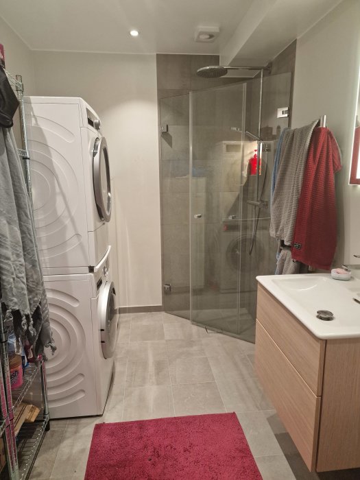 Ett modernt badrum med dusch, tvättmaskin, torktumlare och träfärgad förvaring. Rosa matta på golvet.
