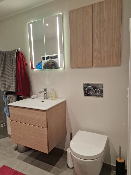 Modernt badrum, träskåp, handfat, toalett, spegel med belysning, grått golv, vit vägg, handdukar.